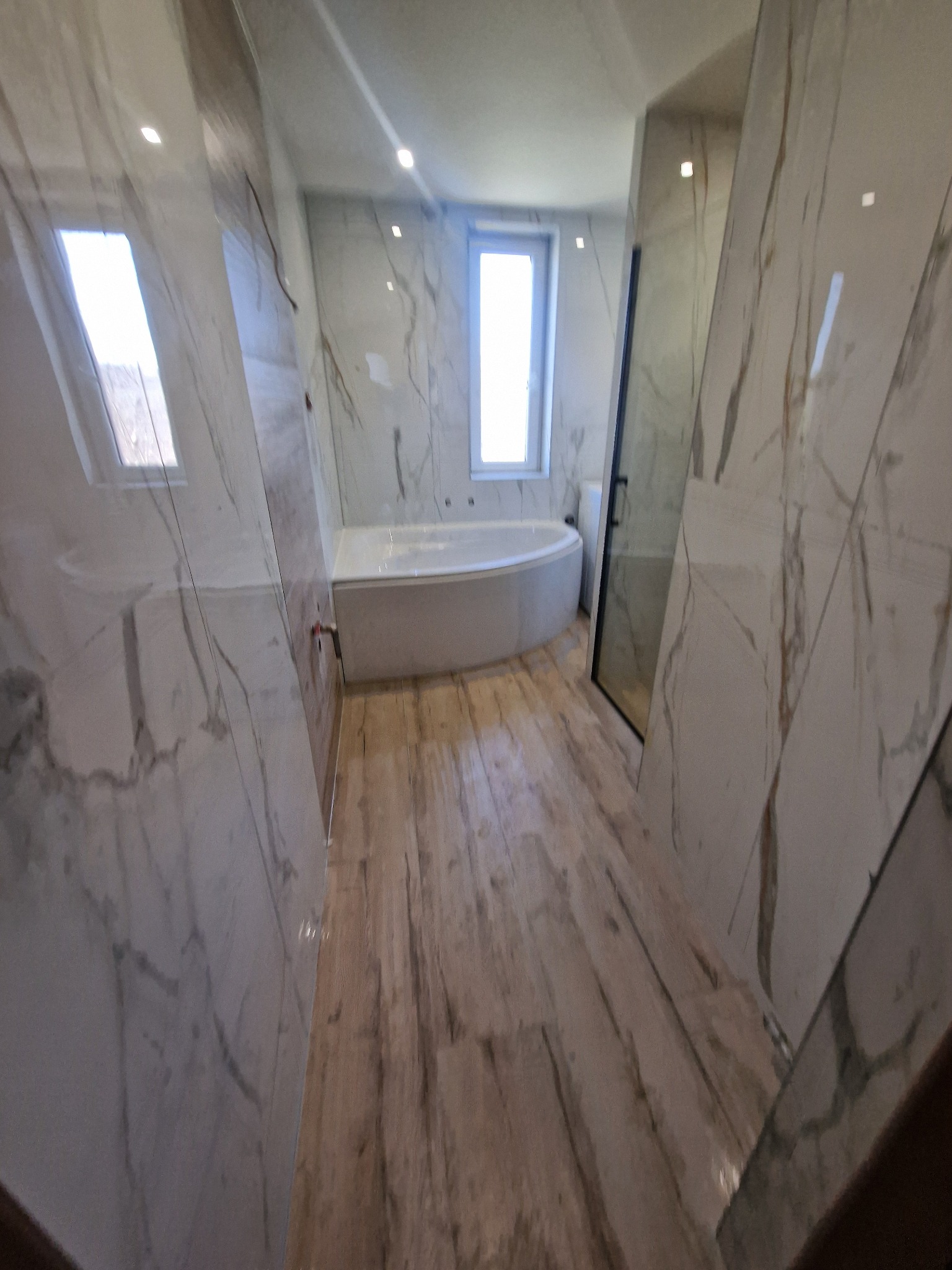 Nowoczesna łazienka z wanną wolnostojącą, kabiną prysznicową z czarnymi profilami i marmurowymi płytkami na ścianach i podłodze imitującej drewno.