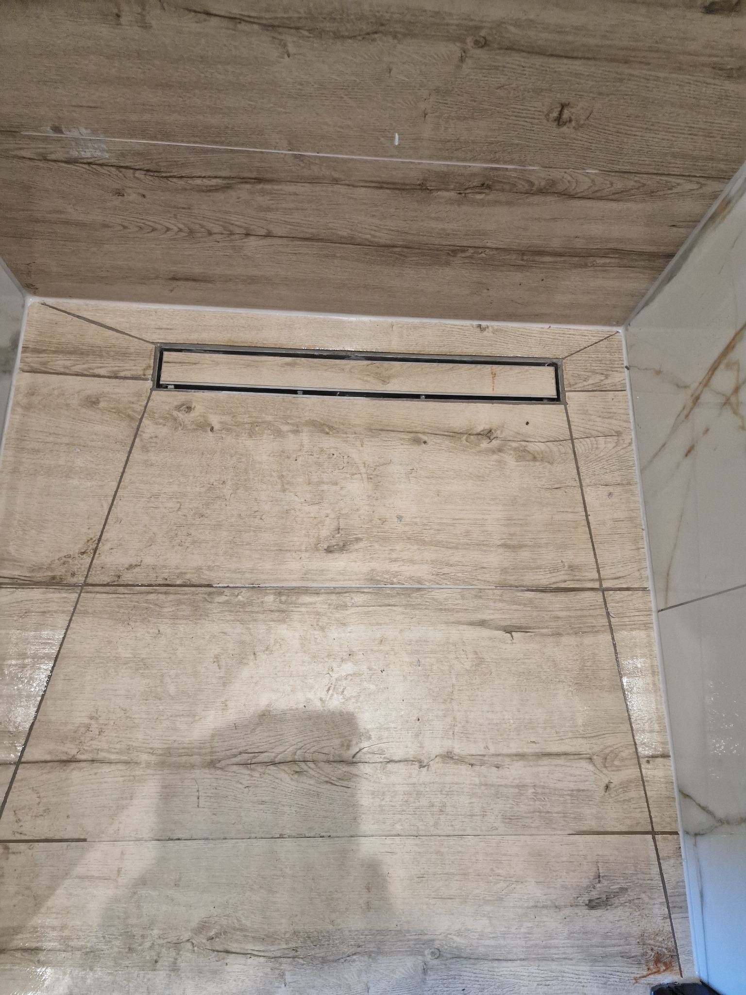 Wykończona kabina prysznicowa z odpływem liniowym, płytki imitujące drewno na podłodze i ścianach, białe płytki marmurowe na ścianie bocznej.