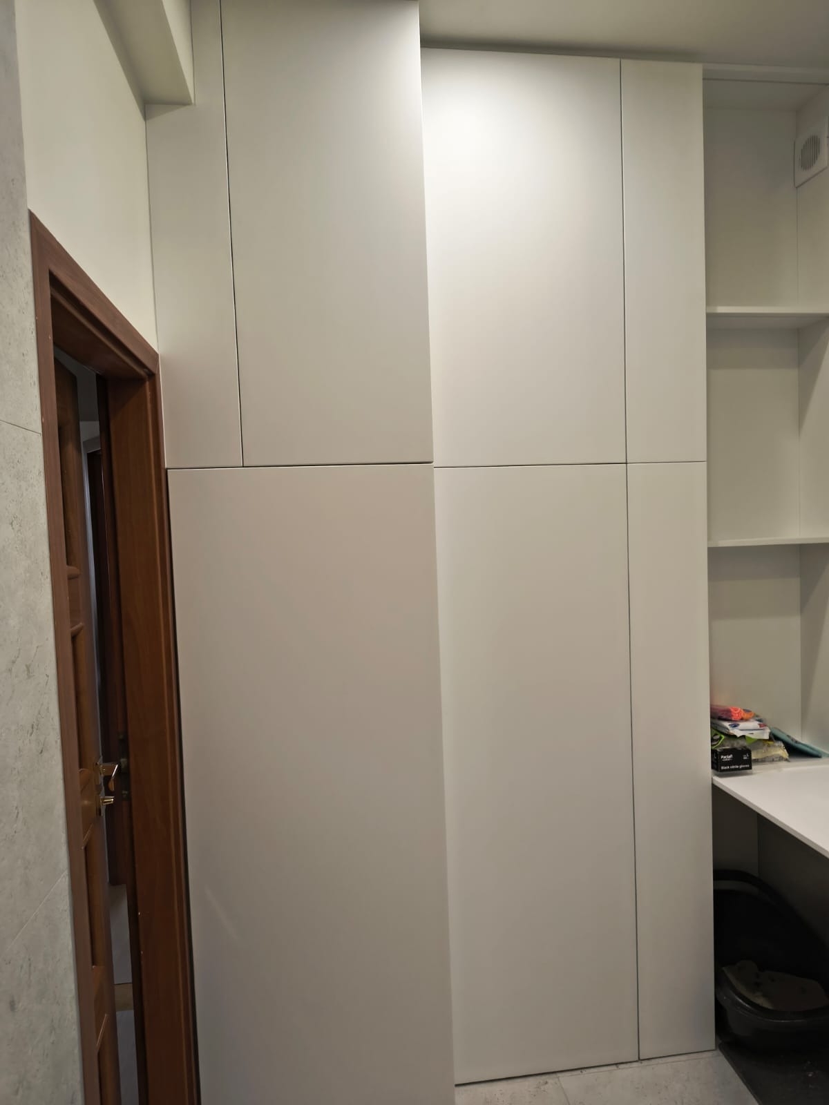 Biała, zabudowana szafa wnękowa z półkami w łazience, widoczny fragment drewnianych drzwi i szara ściana z płytek.