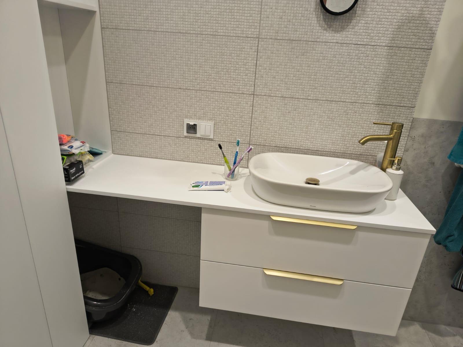 Nowoczesna łazienka z białą umywalką nablatową, złotą baterią i szafką z podwójnymi szufladami oraz mozaiką na ścianie. Widoczne akcesoria łazienkowe: szczoteczki do zębów, pasta do zębów, dozownik...