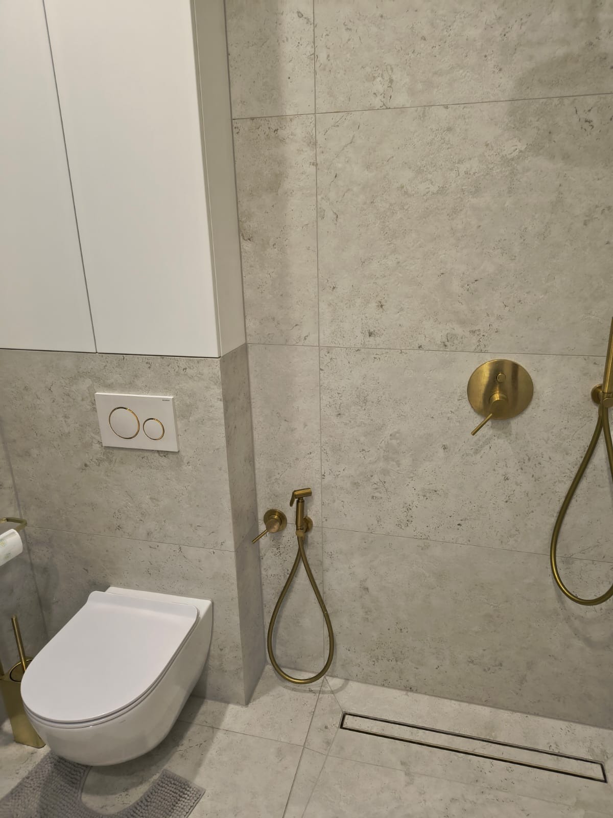 Nowoczesna łazienka z wiszącą toaletą, złotą armaturą i płytkami imitującymi kamień, widoczny odpływ liniowy w podłodze.