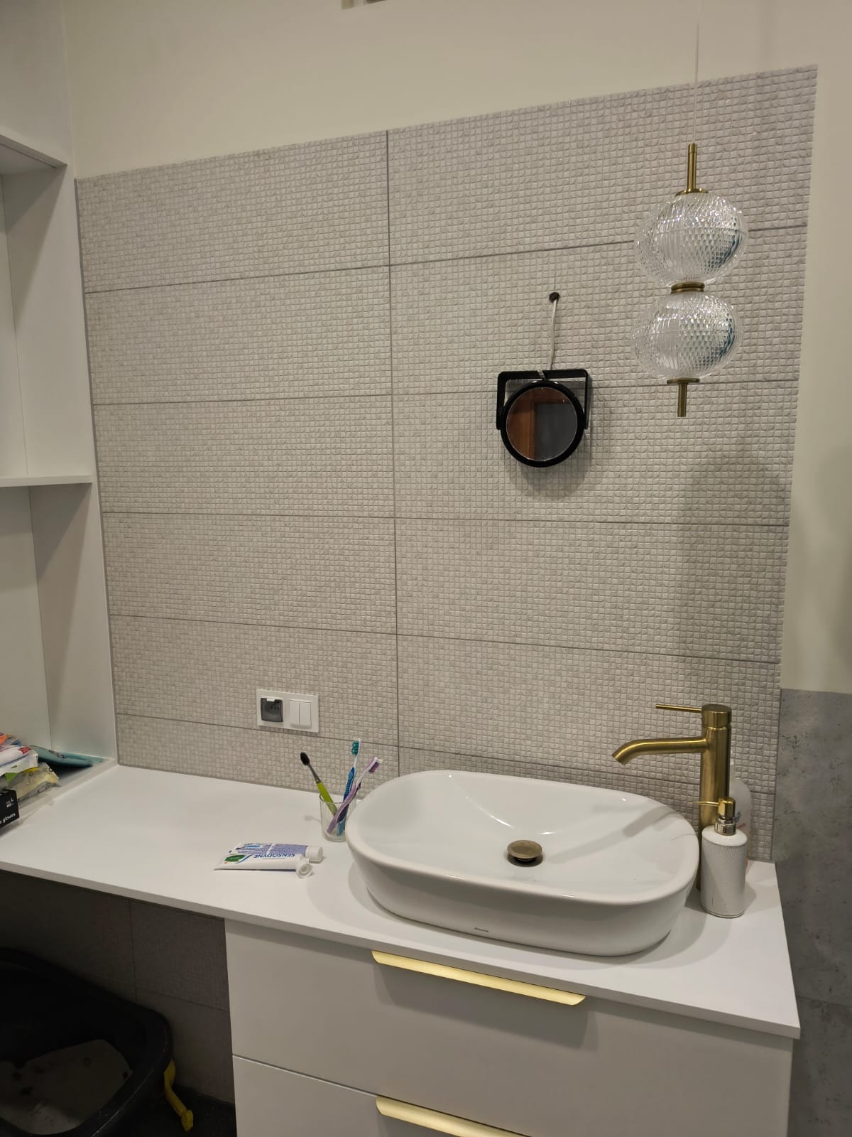 Nowoczesna łazienka z umywalką nablatową, złotą baterią i lustrem na tle ściany wyłożonej szarymi płytkami z delikatną fakturą, wisząca lampa z dwoma szklanymi kloszami.