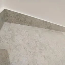 Ukończone połączenie ściany i podłogi z szarej, wzorzystej płytki. Widoczne precyzyjne wykończenie narożnika i fugi. Minimalistyczny design, neutralna kolorystyka.