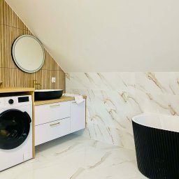 Patryk Machaj - Nowoczesna łazienka na poddaszu z marmurowymi płytkami, czarną wanną i umywalką oraz drewnianymi panelami ściennymi. Widoczna pralka i złota armatura.