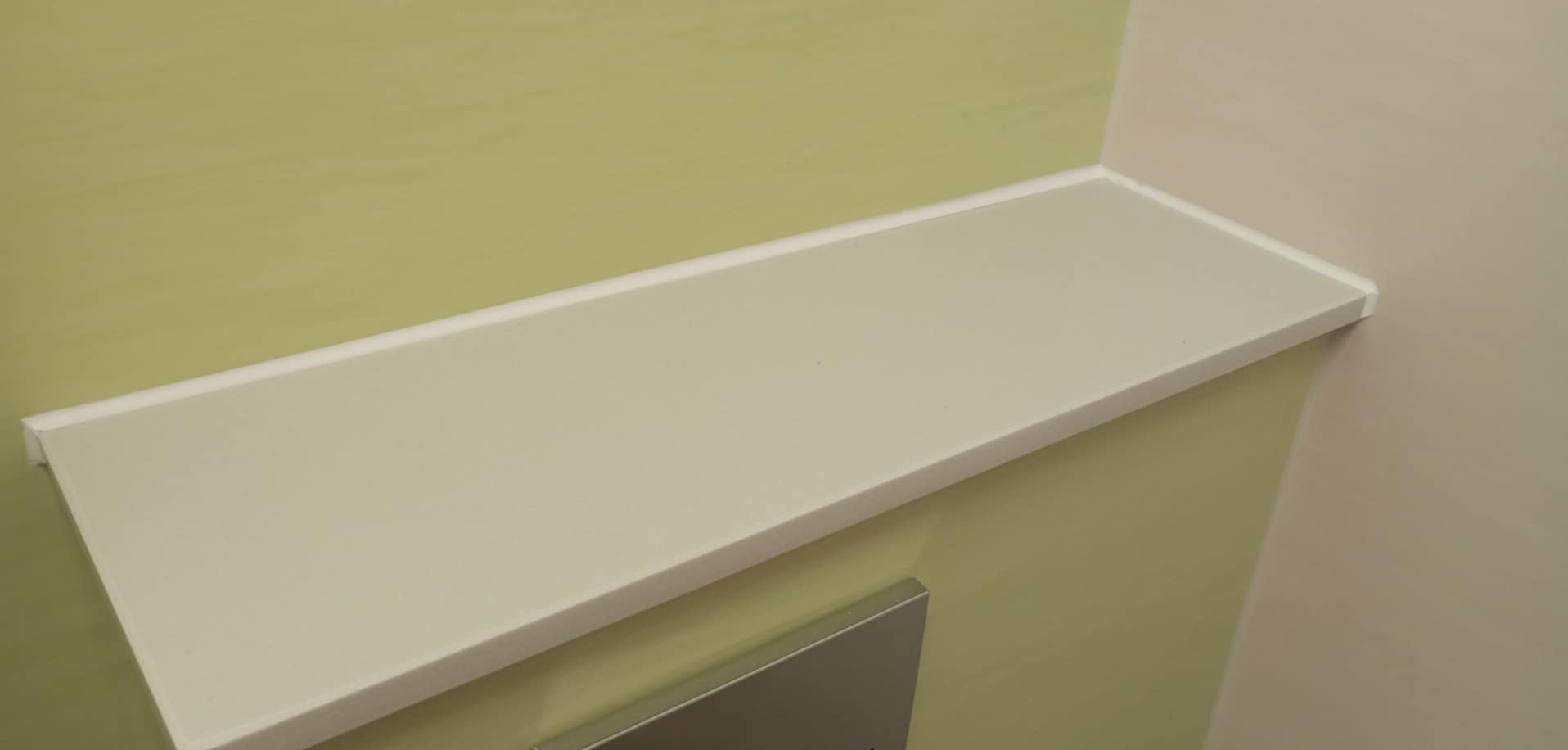 Zamontowana, biała półka w rogu łazienki z zieloną i beżową ścianą, wykończona listwą. Minimalistyczny design, gładka powierzchnia, nowoczesne wykończenie.