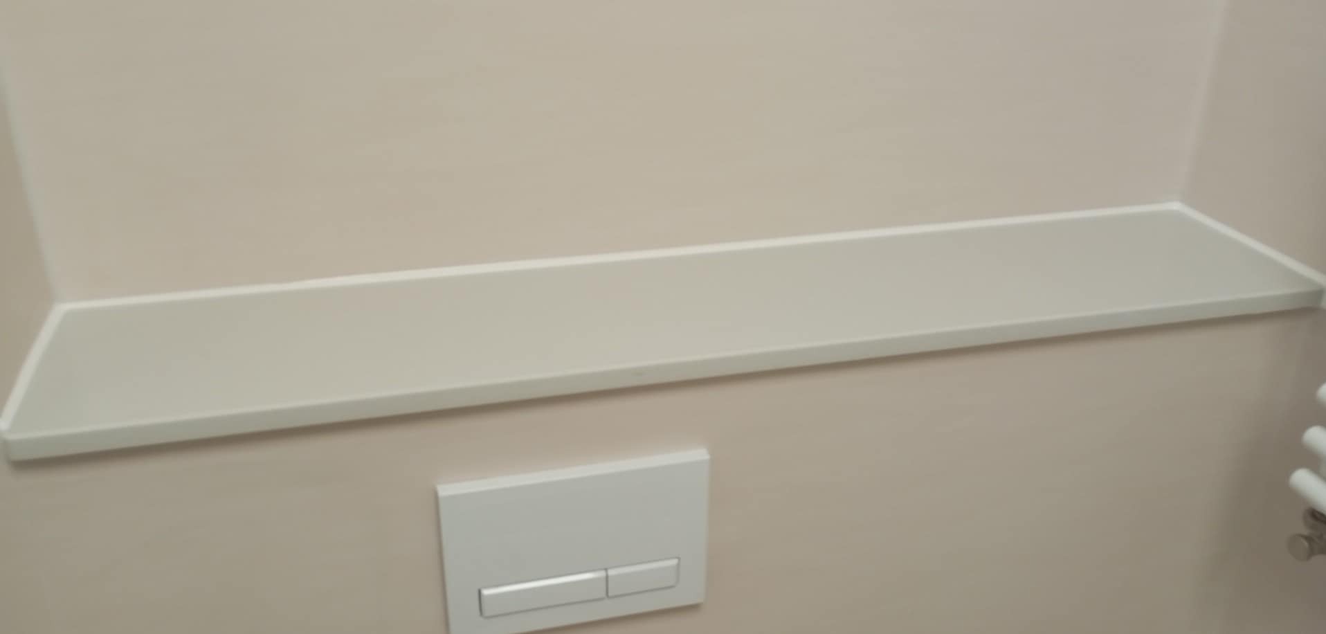 Półka z płytek w łazience nad przyciskiem spłuczki, wykończona w jasnych, neutralnych barwach. Minimalistyczny design, schludne wykonanie.