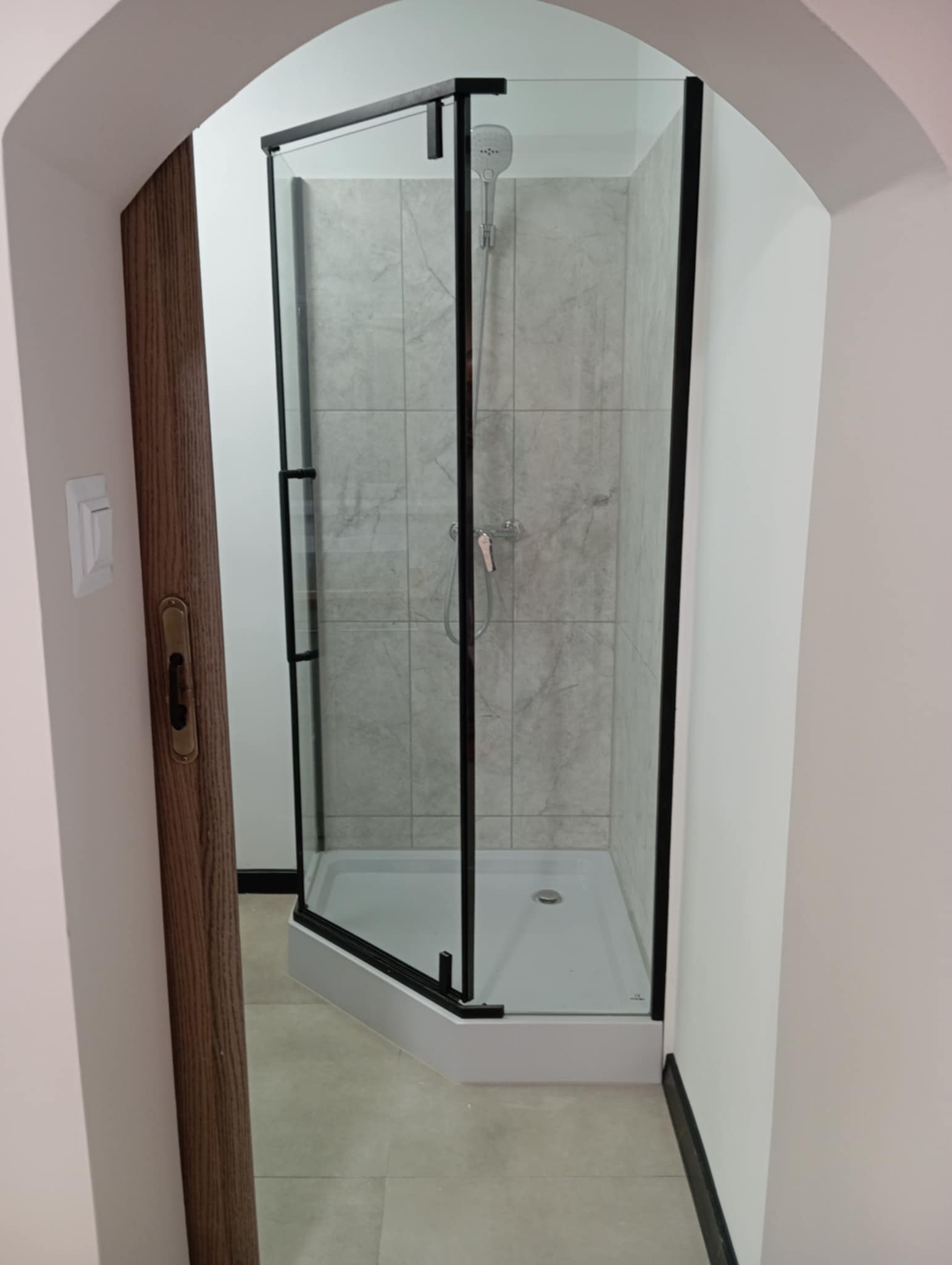 Kabina prysznicowa z czarnymi profilami i szarymi płytkami w łazience, widoczna przez łukowate wejście, z fragmentem drewnianych drzwi po lewej.