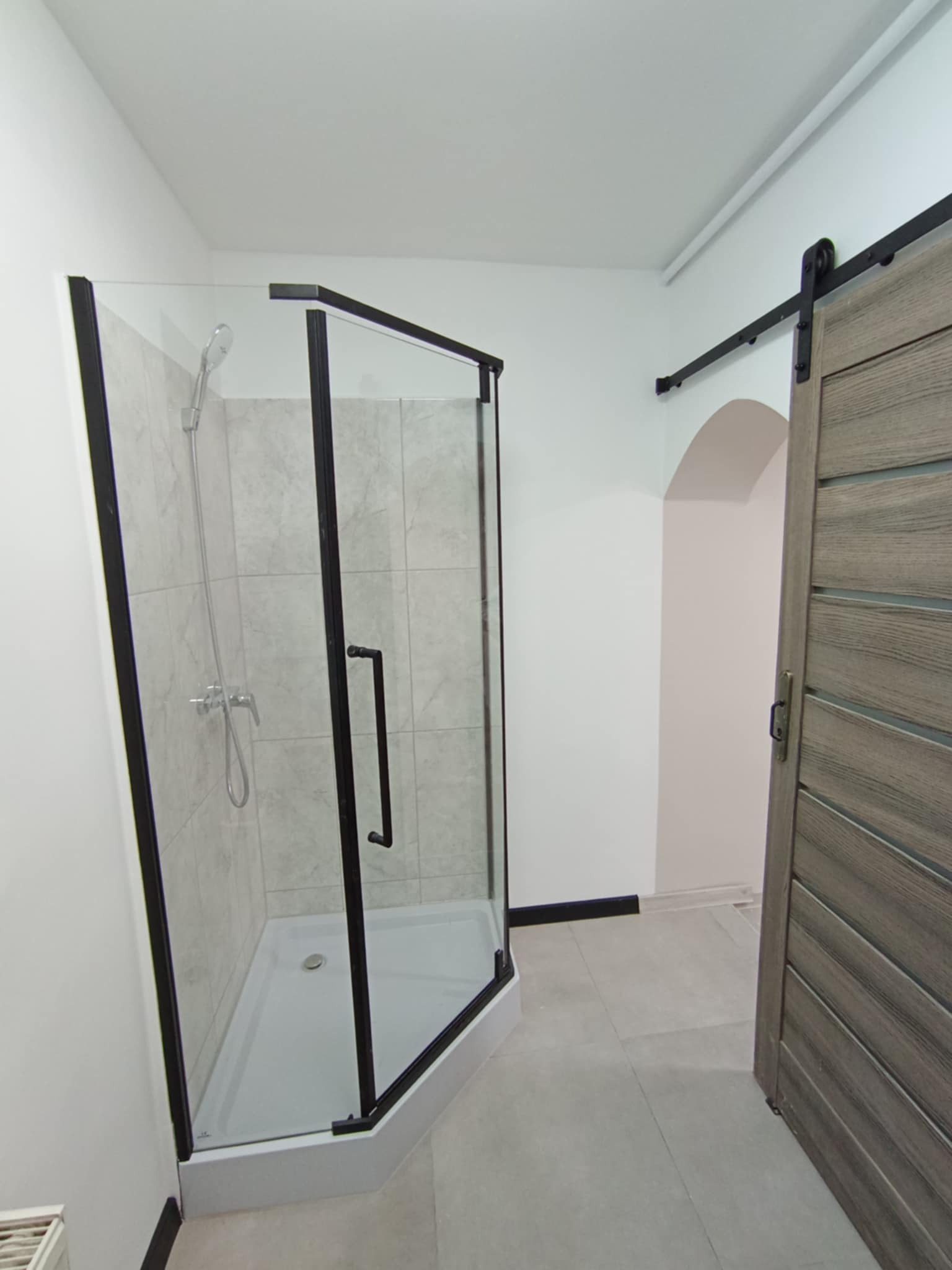 Nowoczesna kabina prysznicowa z czarnymi profilami i przesuwnymi drzwiami w jasnej łazience z szarą podłogą i płytkami. Widoczna drewniana, przesuwna drzwi.