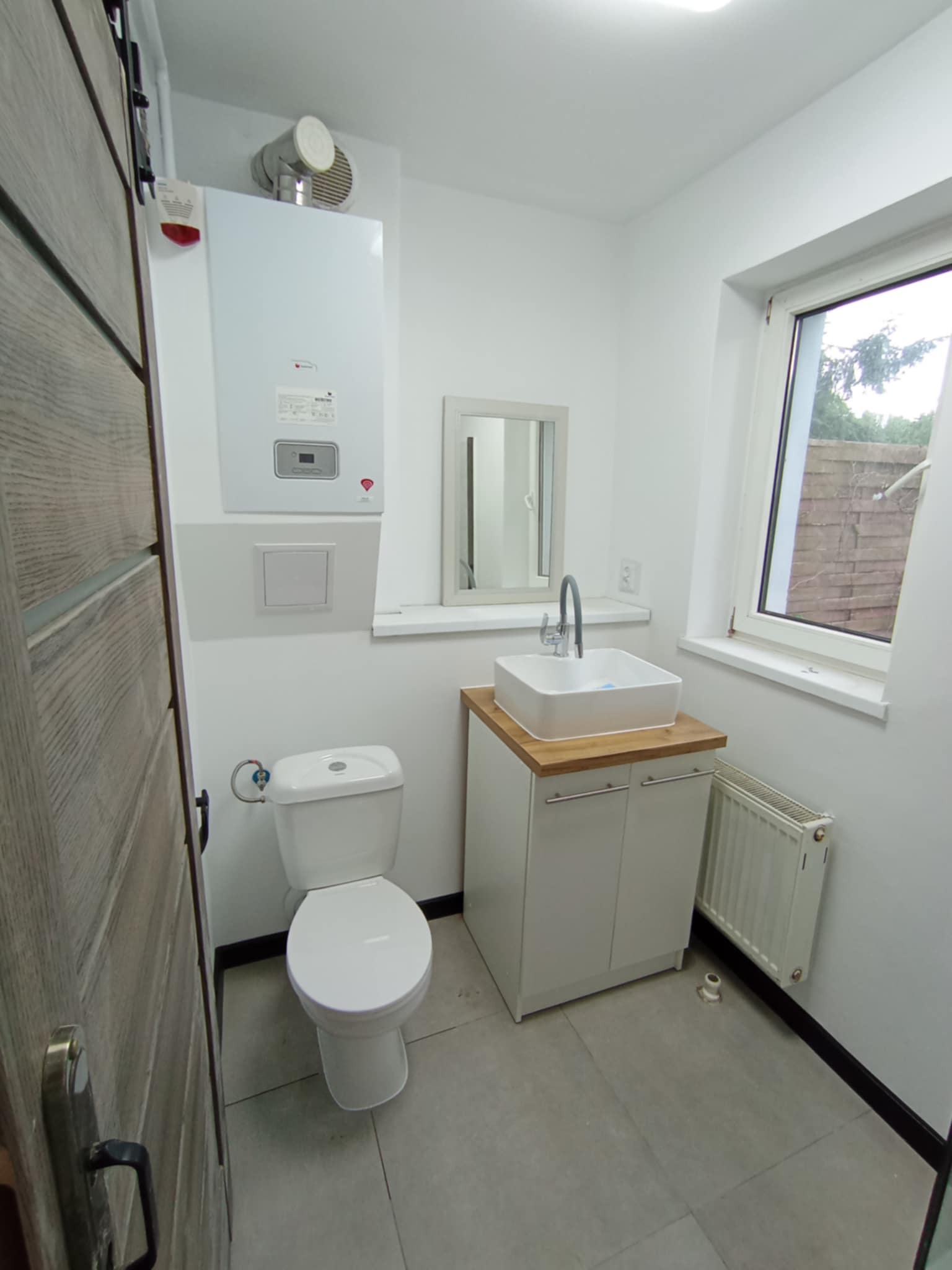 Jasna łazienka po modernizacji: biała armatura, grzejnik, piec gazowy i drewniany blat pod umywalką. Minimalistyczny design z szarymi płytkami podłogowymi.