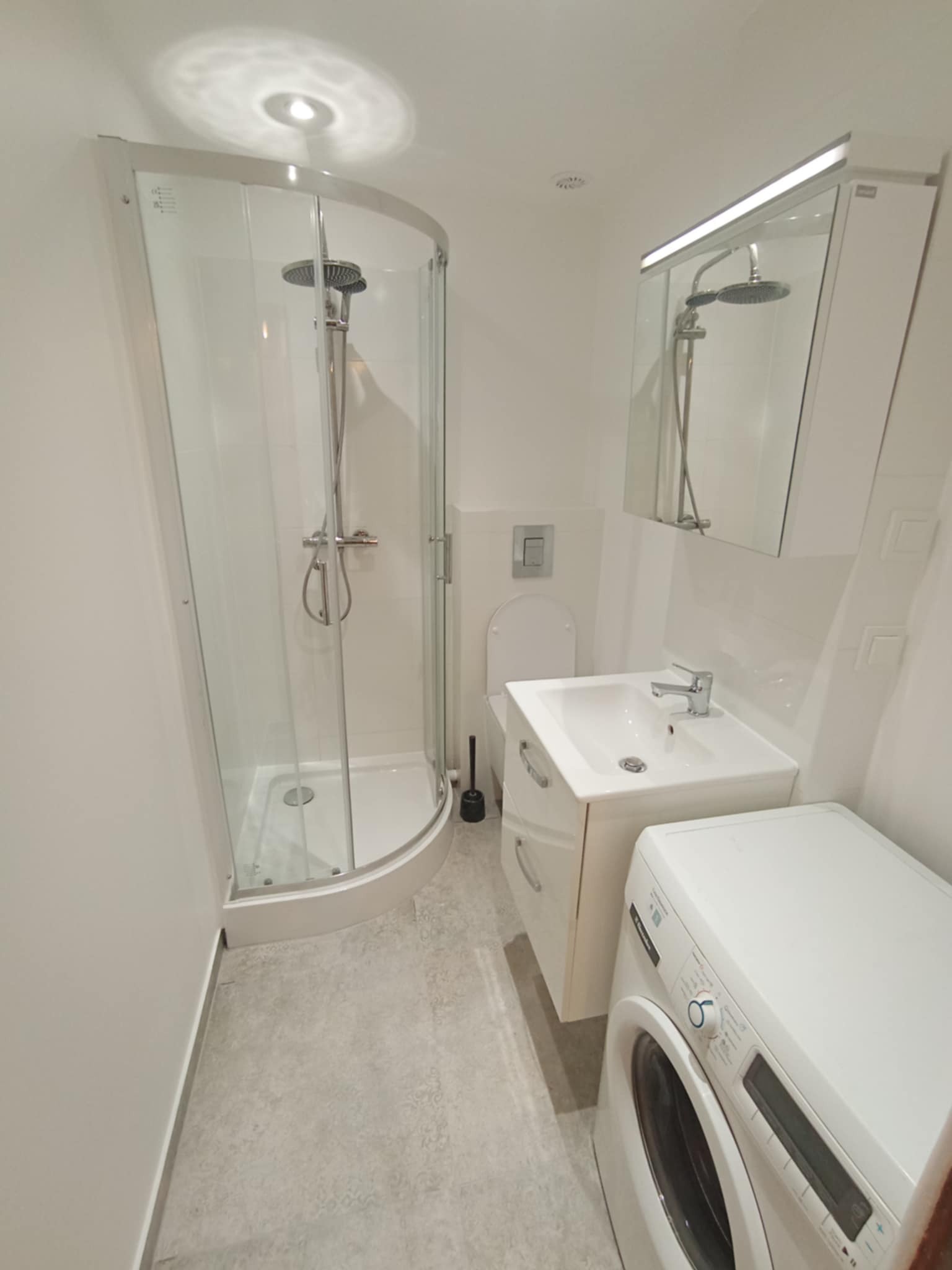 Nowoczesna, jasna łazienka po renowacji z kabiną prysznicową, umywalką z szafką i pralką. Szare płytki podłogowe, białe ściany i armatura.