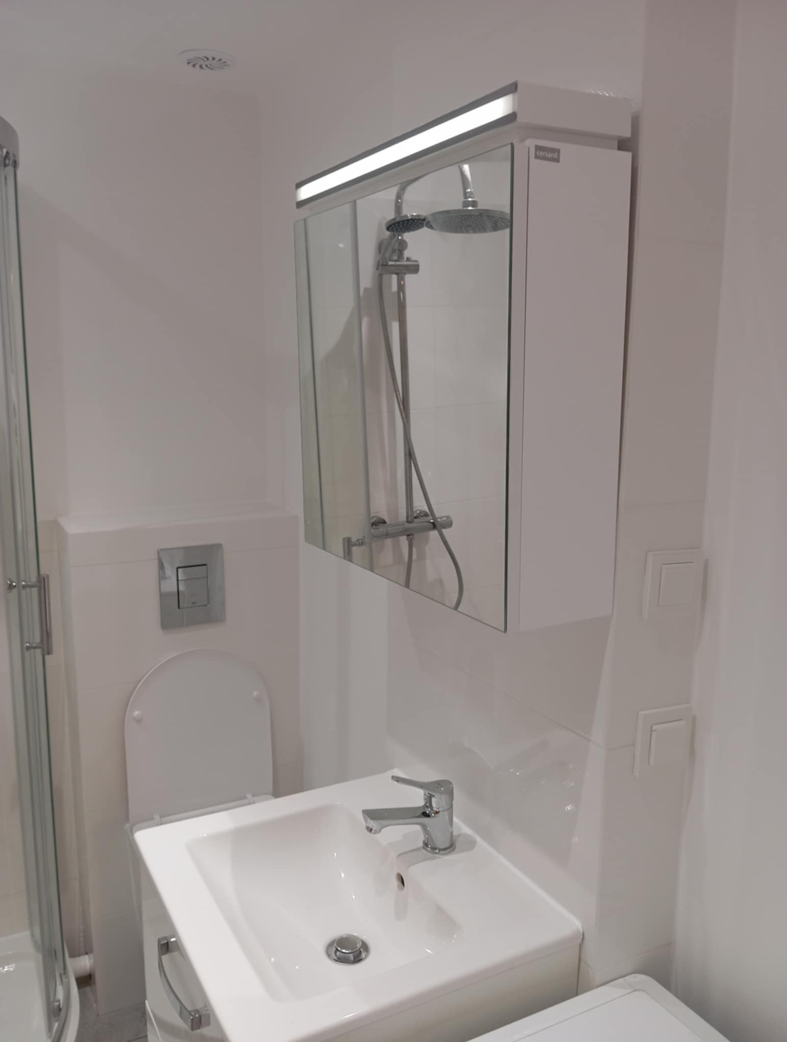 Biała, nowoczesna łazienka po remoncie: umywalka z szafką, toaleta z podtynkową spłuczką i lustro z oświetleniem LED nad kabiną prysznicową.