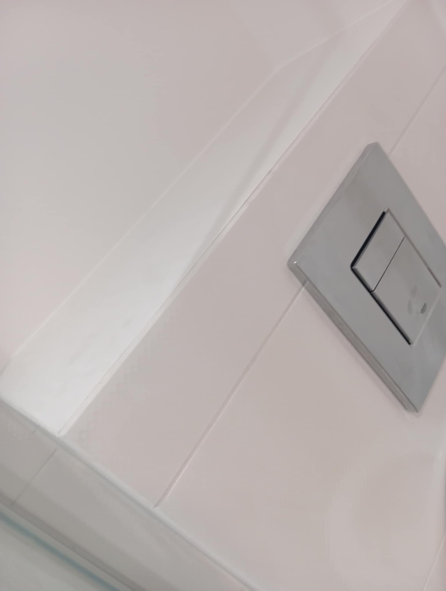 Jasnoróżowe, błyszczące płytki ścienne w łazience z chromowanym przyciskiem spłuczki, ułożone z precyzją i dbałością o detale.