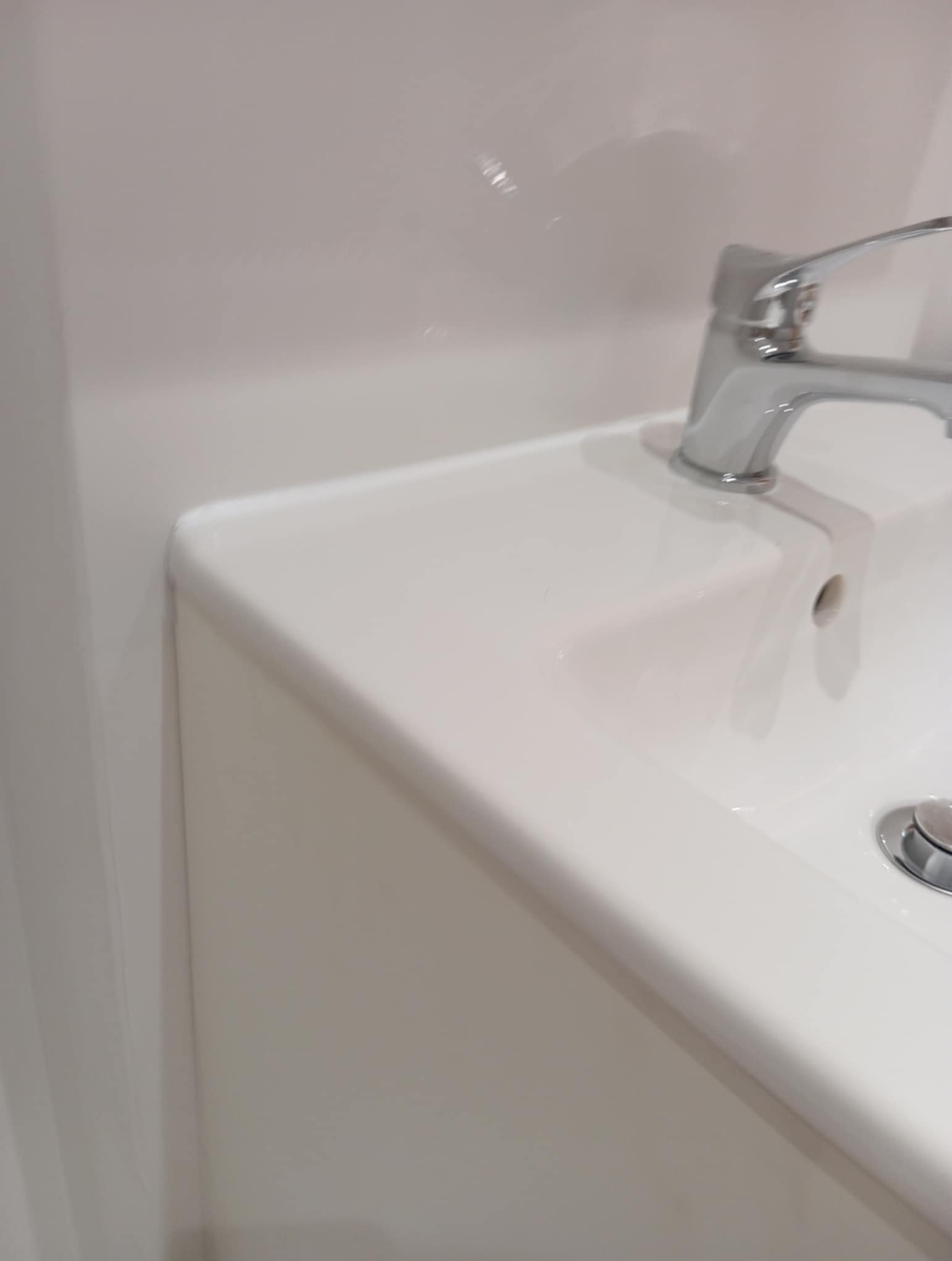 Biała umywalka z chromowaną baterią i odpływem w minimalistycznej łazience. Widoczny fragment ściany. Całość utrzymana w jasnej tonacji.