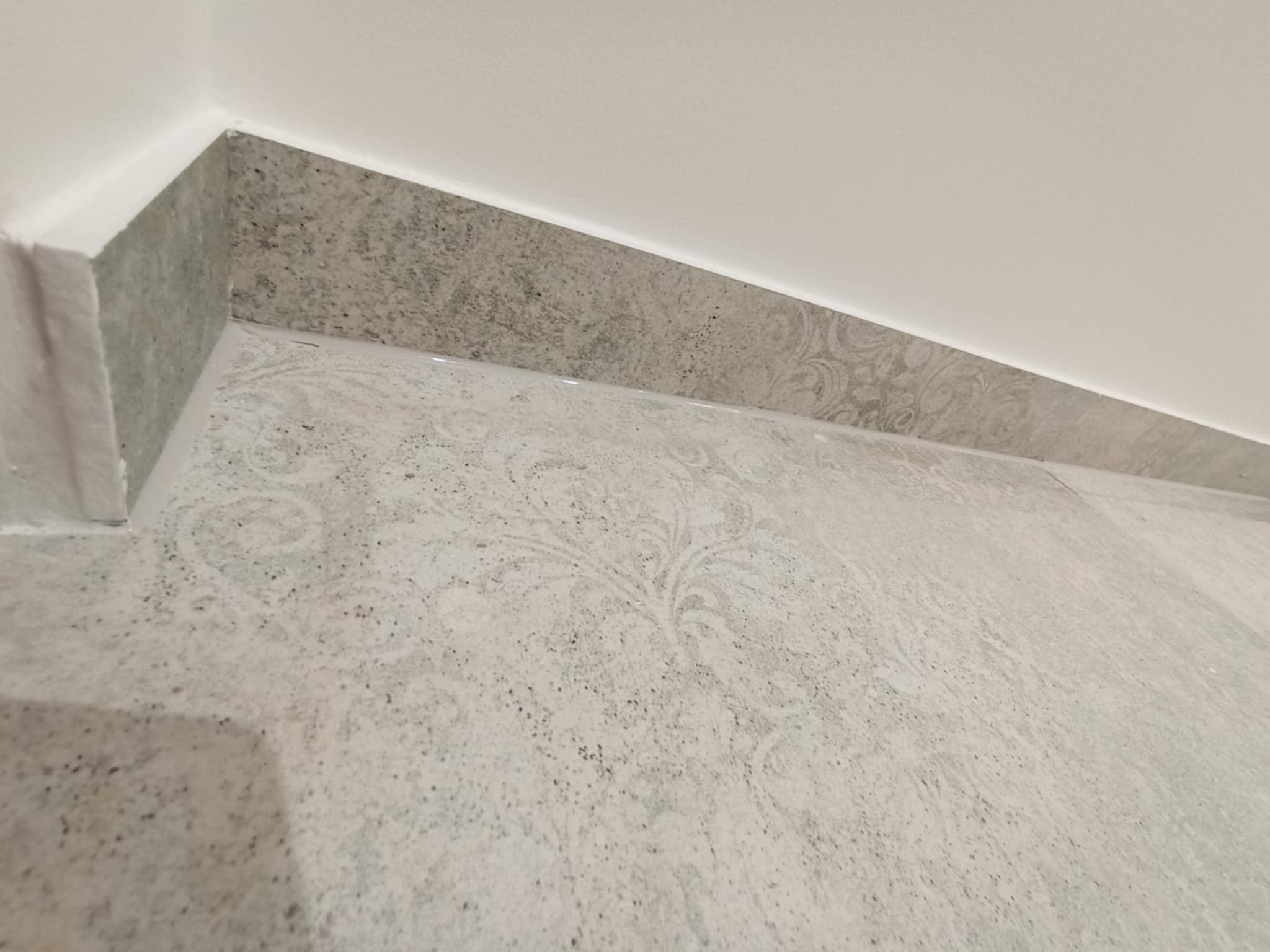 Ukończone połączenie ściany i podłogi z szarej, wzorzystej płytki. Widoczne precyzyjne wykończenie narożnika i fugi. Minimalistyczny design, neutralna kolorystyka.