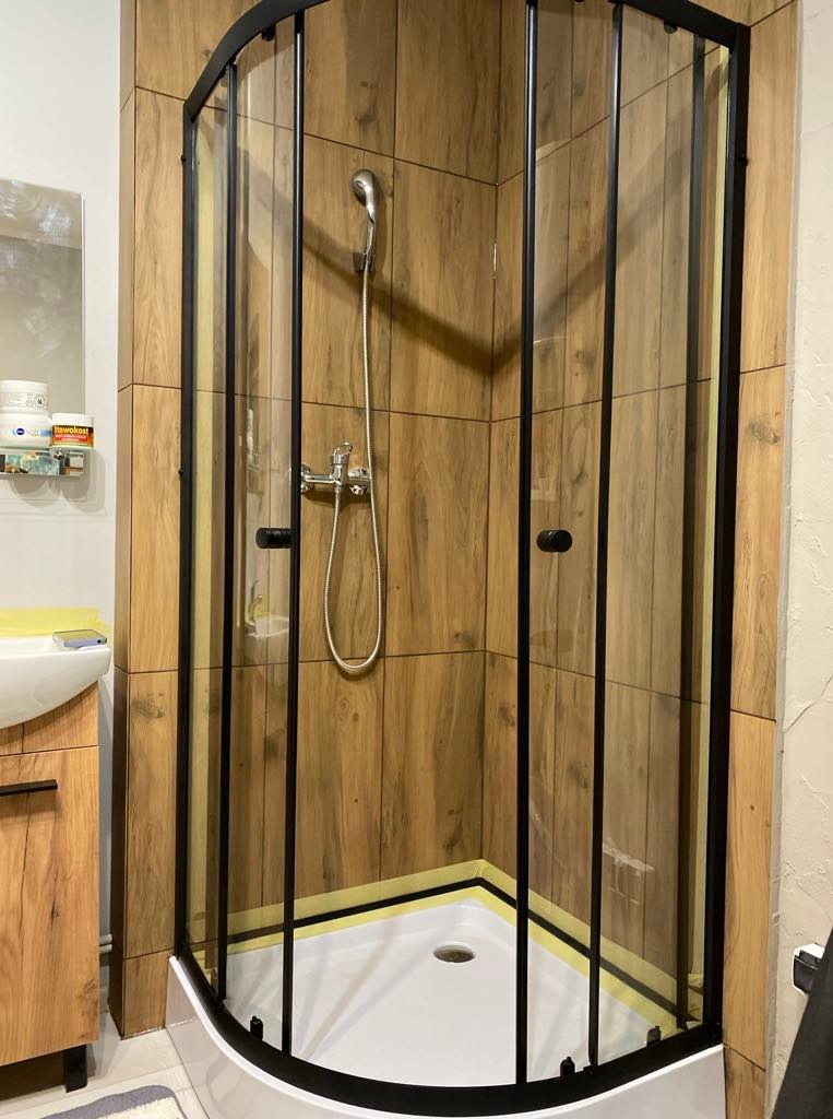 Kabina prysznicowa z czarnymi profilami i drewnopodobnymi płytkami w Radowo Małe. Nowoczesna aranżacja łazienki z umywalką i szafką w tle.