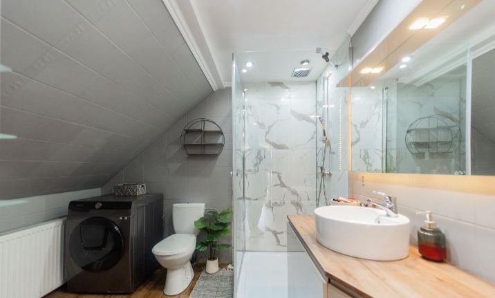 Nowoczesna łazienka na poddaszu ze skośnym sufitem, kabiną prysznicową ze szklanymi drzwiami i pralką w ciemnej obudowie. Marmurowe płytki na ścianach.