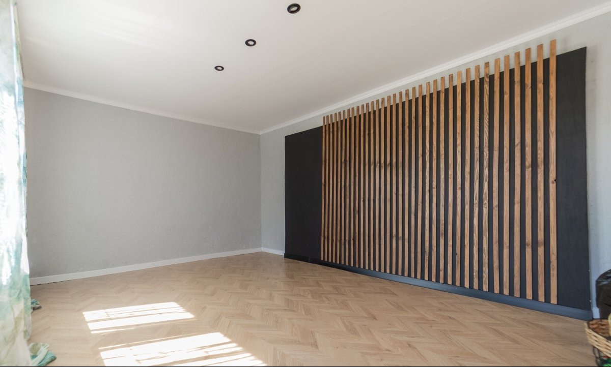 Jasne wnętrze po renowacji z drewnianą ścianką działową na czarnym tle i parkietem w jodełkę. Widoczne oświetlenie sufitowe.