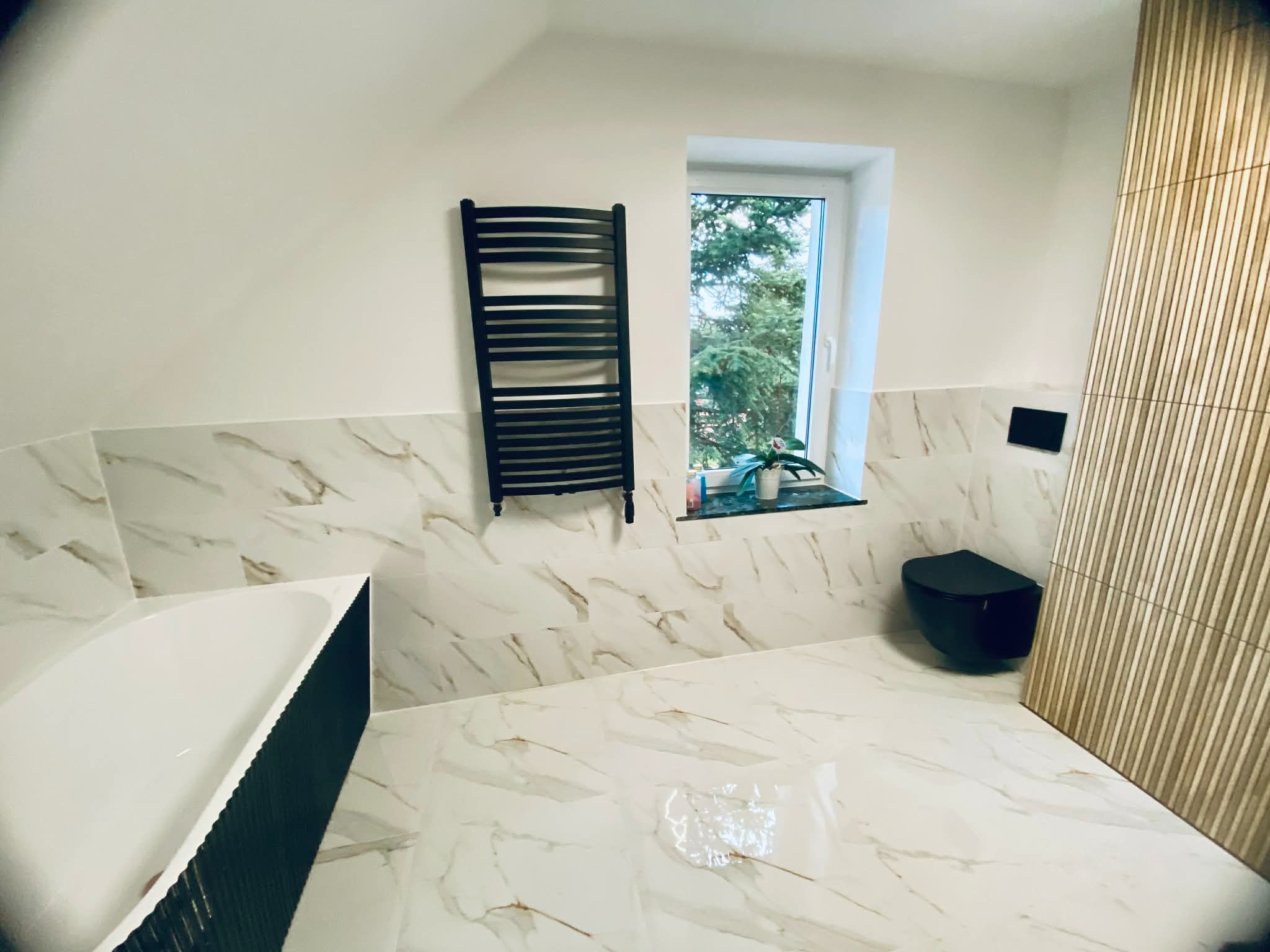 Wykończona łazienka na poddaszu z marmurowymi płytkami, czarną armaturą i grzejnikiem drabinkowym. Widok na okno z zielenią. Nowoczesny design.
