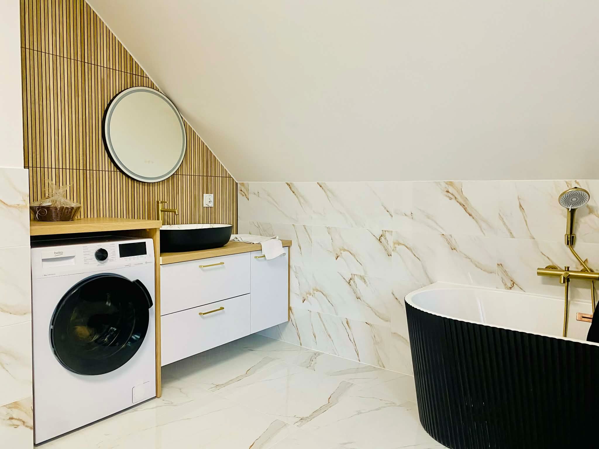 Nowoczesna łazienka na poddaszu z marmurowymi płytkami, czarną wanną i umywalką oraz drewnianymi panelami ściennymi. Widoczna pralka i złota armatura.