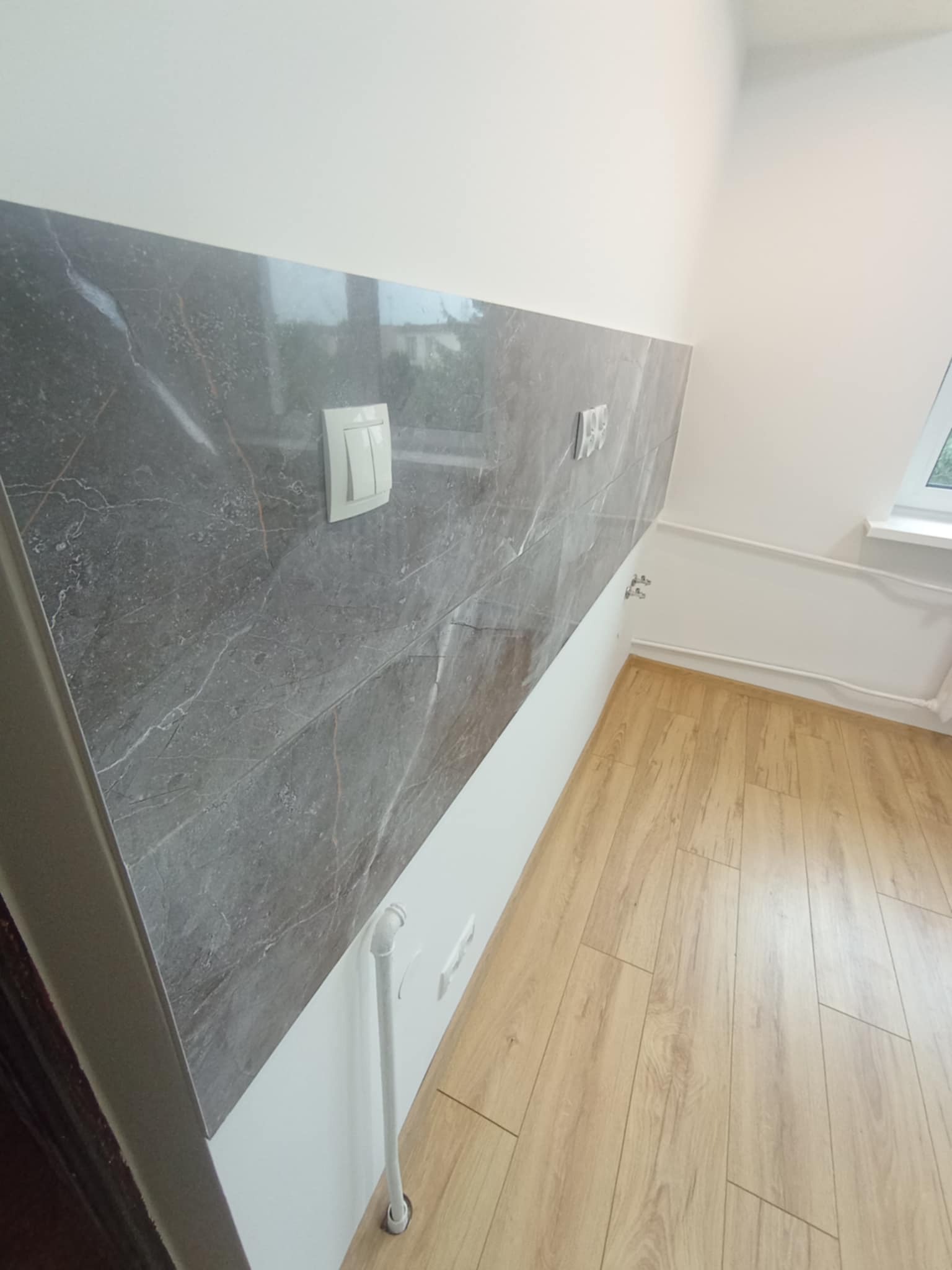 Wykończona ściana z szarych płytek imitujących marmur, z zamontowanym włącznikiem światła. Na podłodze panele w odcieniach drewna. Białe ściany i listwy przypodłogowe.