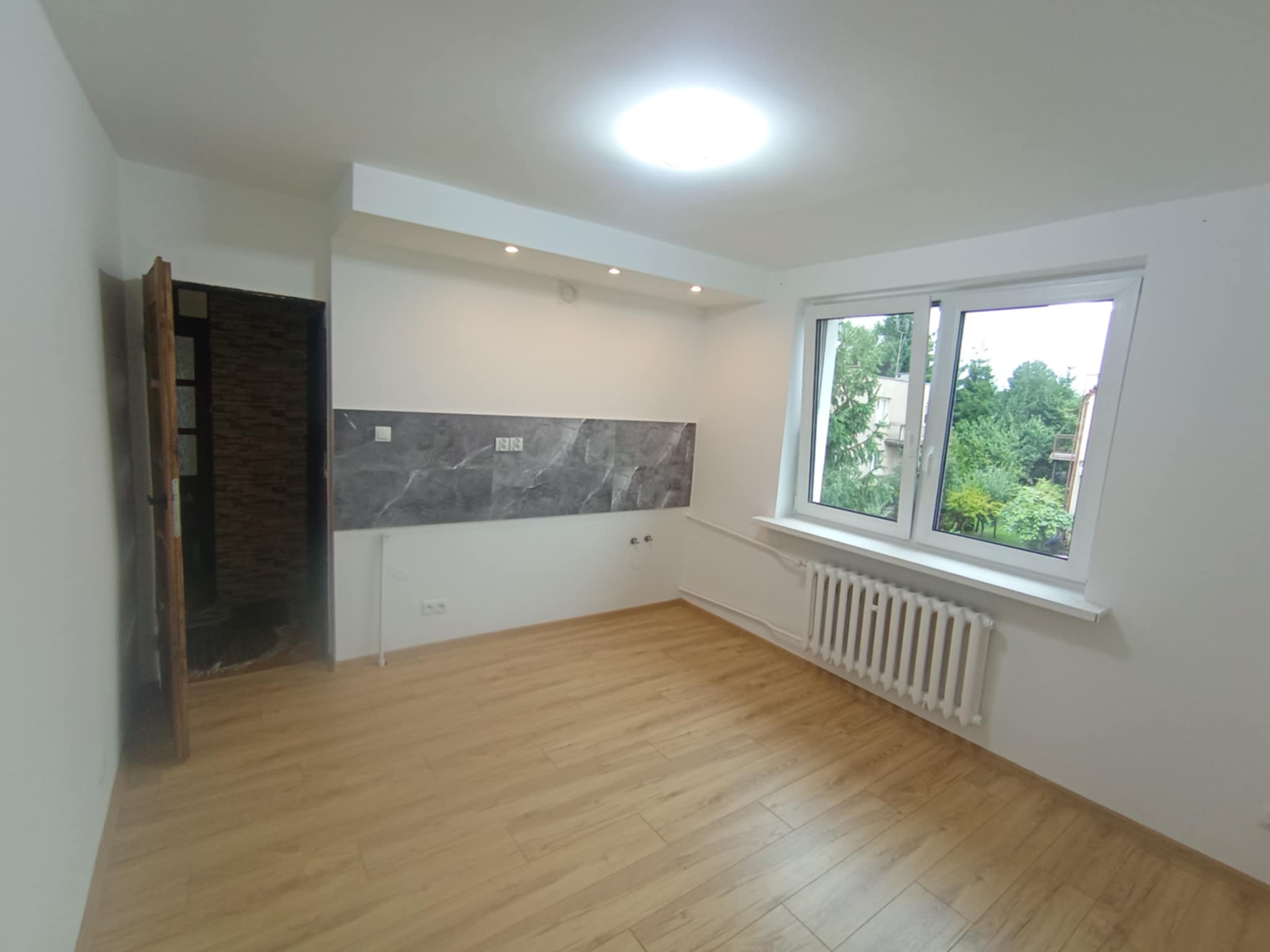 Jasne wnętrze po remoncie: drewnopodobna podłoga, białe ściany, okno z widokiem na zieleń, szary panel dekoracyjny na ścianie. Otwór drzwiowy bez drzwi.