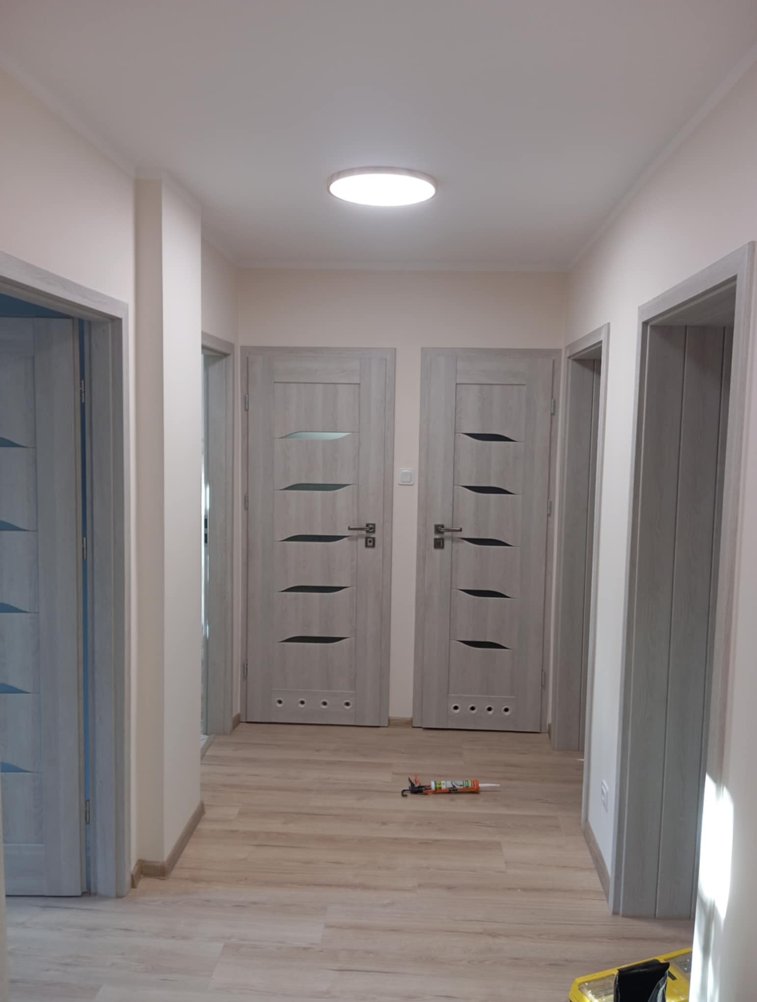 Jasny korytarz z trzema parami drzwi w odcieniach szarości, z widoczną tubą silikonu na podłodze i lampą sufitową. Widok na wykończone wnętrze.