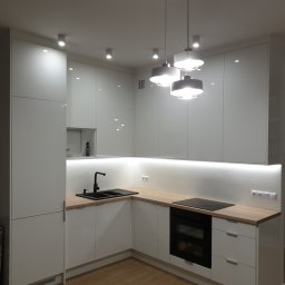 NINTA.PL - Nowoczesna, biała kuchnia z drewnianym blatem, czarnym zlewem i płytą indukcyjną, oświetlona lampami wiszącymi i listwą LED pod szafkami.