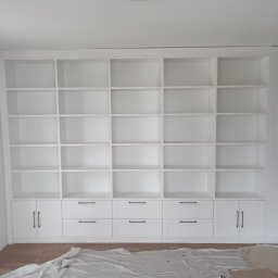 NINTA.PL - Biała szafa wnękowa z licznymi półkami i szufladami z czarnymi uchwytami, zabezpieczona folią malarską na podłodze, widok z dołu.