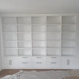 Biała szafa wnękowa z licznymi półkami i szufladami z czarnymi uchwytami, zabezpieczona folią malarską na podłodze, widok z dołu.