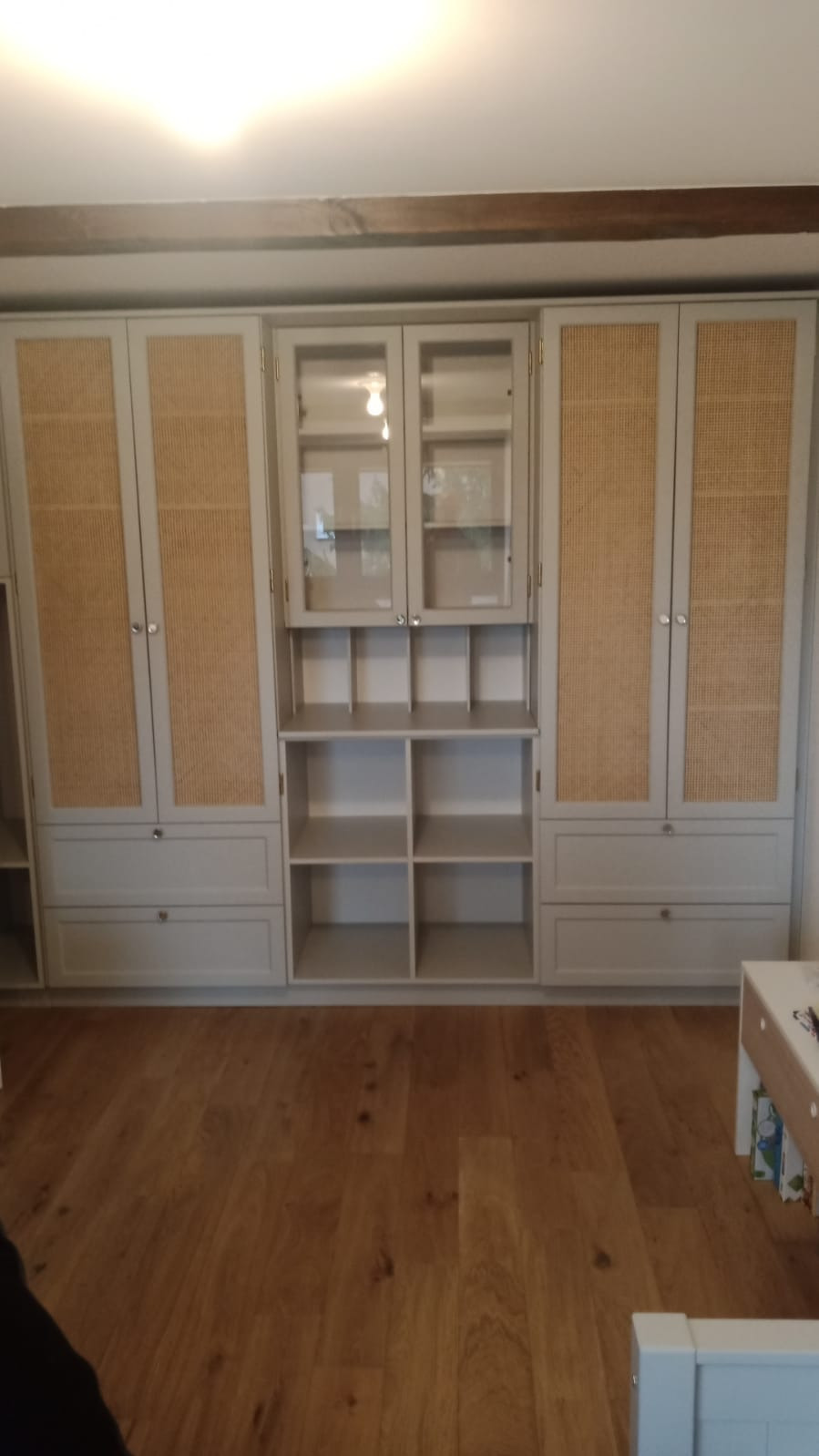 Szara zabudowa meblowa z rattanowymi frontami, przeszklona witryna i otwarte półki, drewniana podłoga.