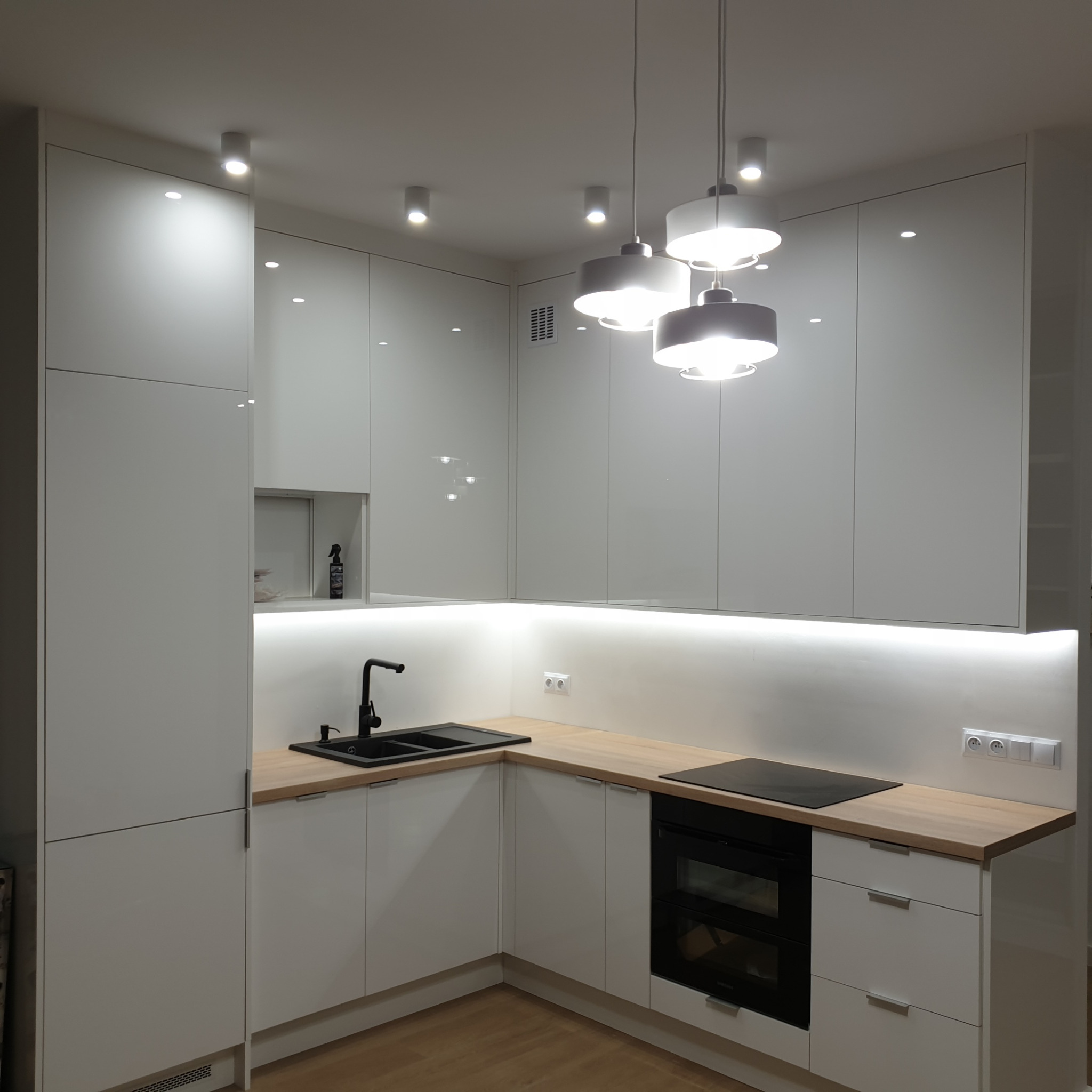 Nowoczesna, biała kuchnia z drewnianym blatem, czarnym zlewem i płytą indukcyjną, oświetlona lampami wiszącymi i listwą LED pod szafkami.