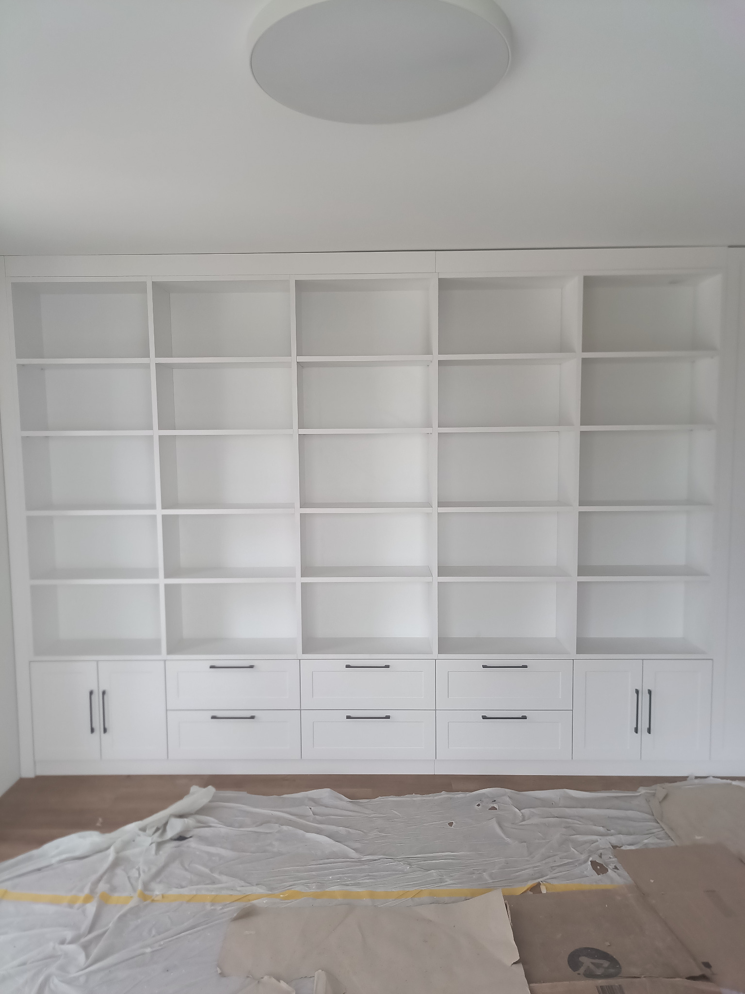 Biała szafa wnękowa z licznymi półkami i szufladami z czarnymi uchwytami, zabezpieczona folią malarską na podłodze, widok z dołu.