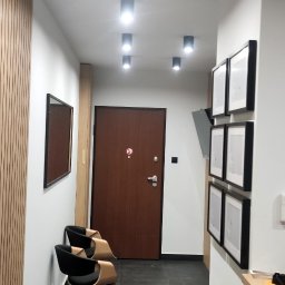 ŁUKASZ MOŹDZIERZ - Nowoczesny korytarz w Krakowie z szarą podłogą, drewnianą ścianą z listew, rzędem okrągłych lamp sufitowych, brązowymi drzwiami i dwoma krzesłami biurowymi.