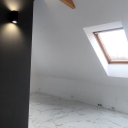 ŁUKASZ MOŹDZIERZ - Wykończone poddasze z drewnianymi belkami, czarnym oświetleniem punktowym, białymi ścianami i podłogą z płytek imitujących marmur.