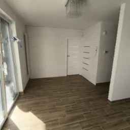 Jasne wnętrze po remoncie z drewnopodobną podłogą z płytek, ścianą z białej cegły i nowoczesnym żyrandolem kryształowym.