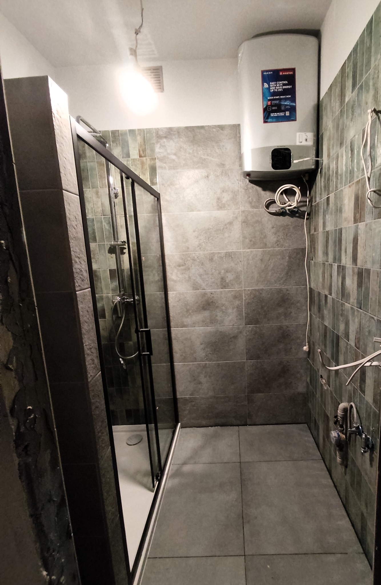 Nowoczesna łazienka z kabiną prysznicową typu walk-in z czarnymi profilami, płytkami w odcieniach szarości i zieleni, oraz podgrzewaczem wody Ariston z widocznymi przewodami.