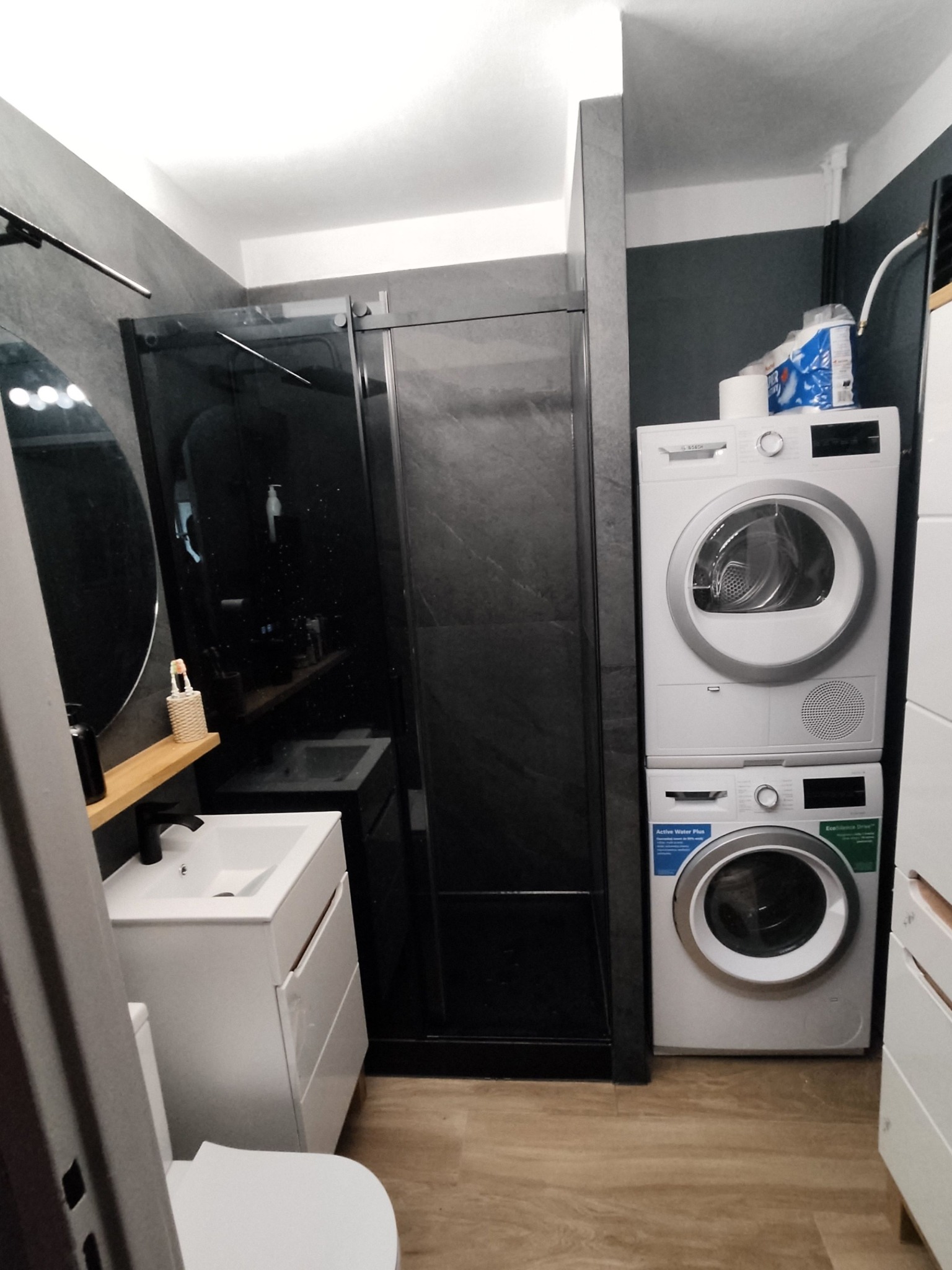 Nowoczesna łazienka z kabiną prysznicową typu walk-in z czarnymi elementami, białą umywalką z szafką, okrągłym lustrem i pralko-suszarką ustawioną jedna na drugiej.