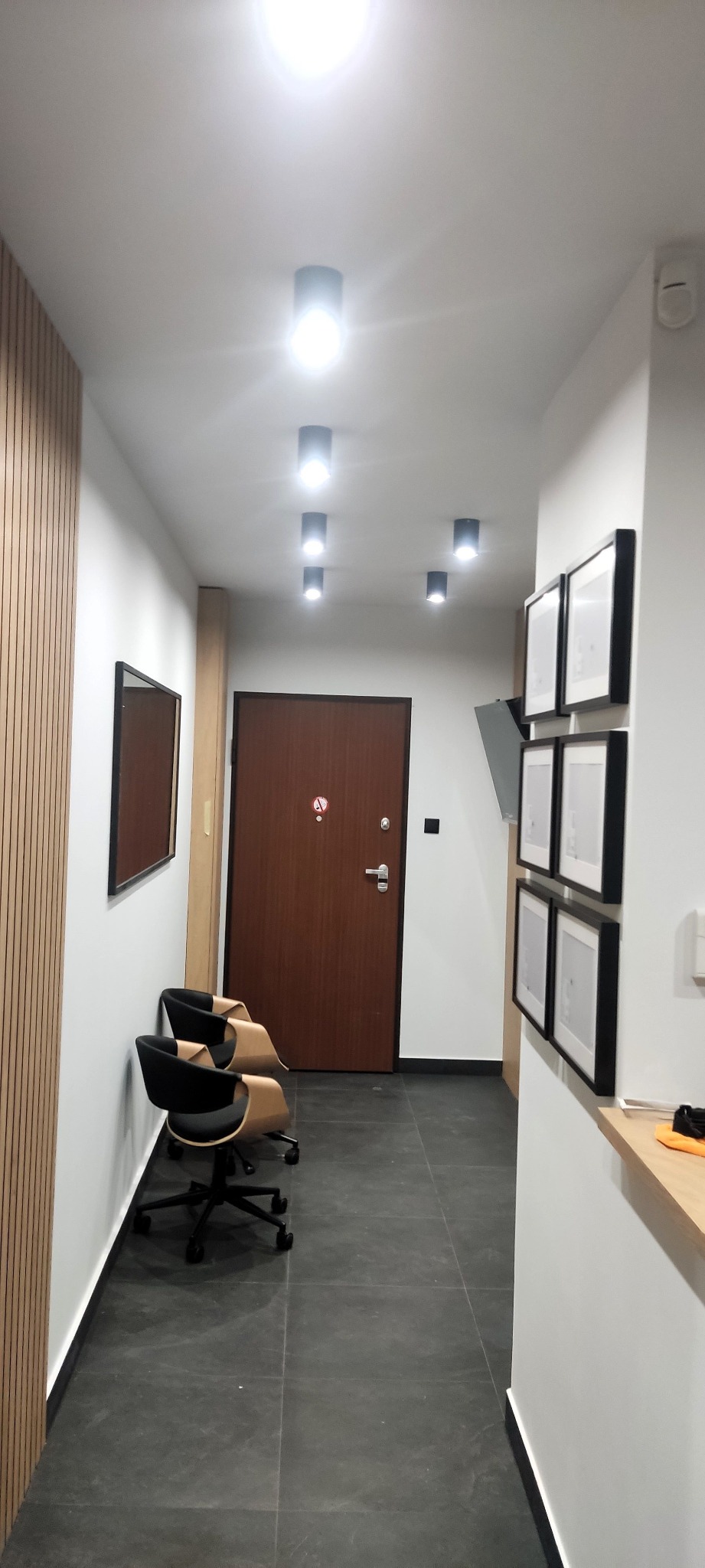 Nowoczesny korytarz w Krakowie z szarą podłogą, drewnianą ścianą z listew, rzędem okrągłych lamp sufitowych, brązowymi drzwiami i dwoma krzesłami biurowymi.