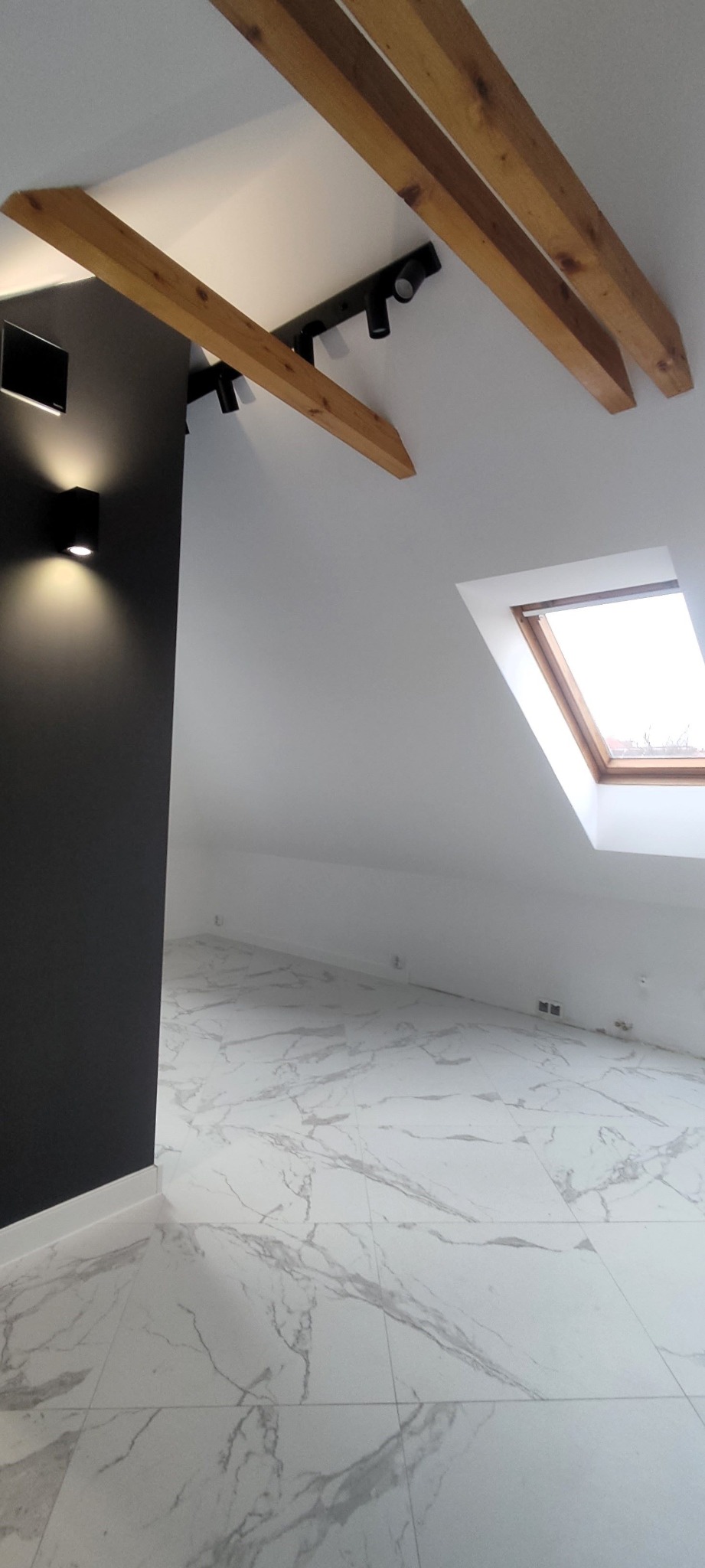 Wykończone poddasze z drewnianymi belkami, czarnym oświetleniem punktowym, białymi ścianami i podłogą z płytek imitujących marmur.