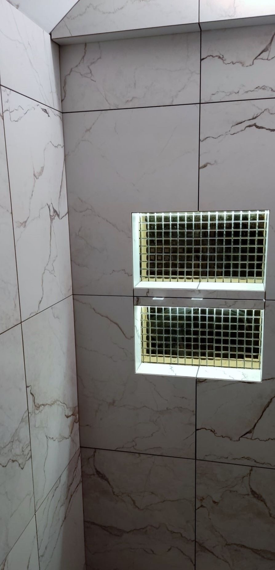 Wykończona ściana łazienki z białych płytek imitujących marmur i podświetlanymi, mozaikowymi wnękami.