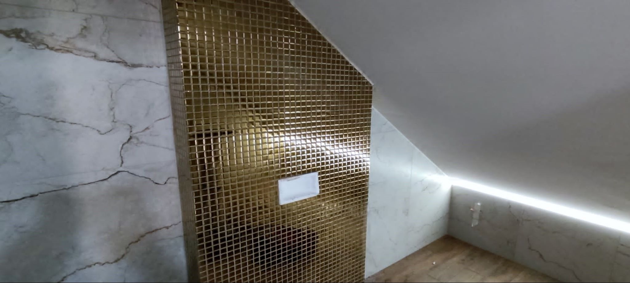 Wykończona łazienka na poddaszu z błyszczącą, złotą mozaiką i marmurowymi płytkami, oświetlona listwą LED.