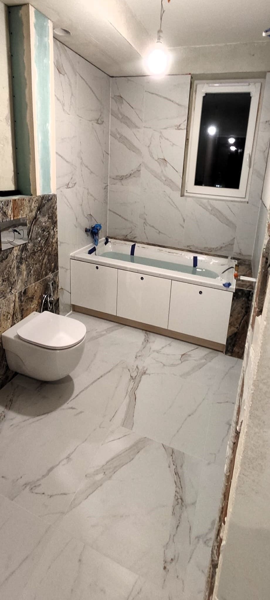Wnętrze łazienki w trakcie prac wykończeniowych: zamontowana podwieszana toaleta, obudowana wanna z wypełnioną wodą, marmurowe płytki na ścianach i podłodze, widoczne niedokończone fragmenty ścian.