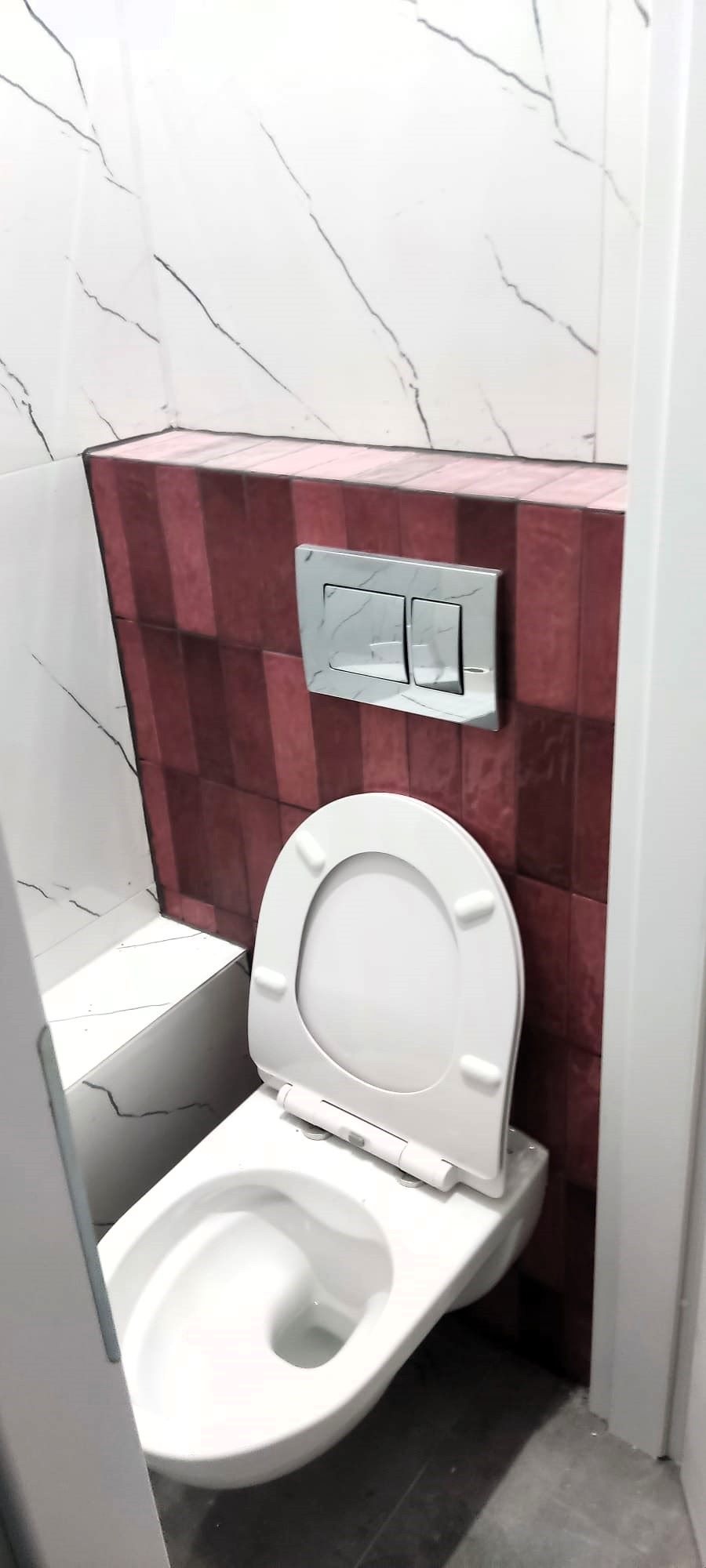 Nowoczesna toaleta podwieszana z białym sedesem i marmurowym przyciskiem spłukującym na tle bordowej ściany z kwadratowych płytek, uzupełniona białymi płytkami imitującymi marmur.