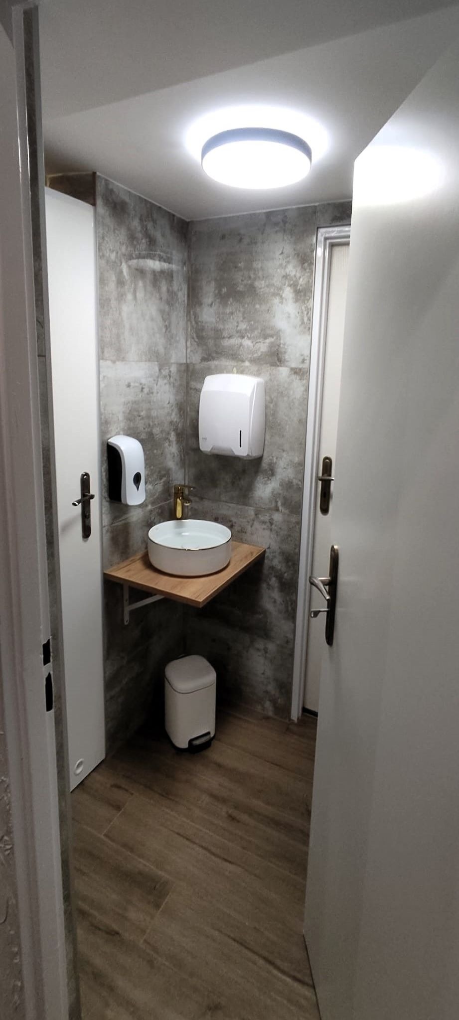 Nowoczesna, mała łazienka z okrągłą umywalką nablatową na drewnianym blacie, szare płytki imitujące beton, białe drzwi i podajniki na mydło oraz papier, drewnopodobna podłoga i okrągła lampa sufitowa.