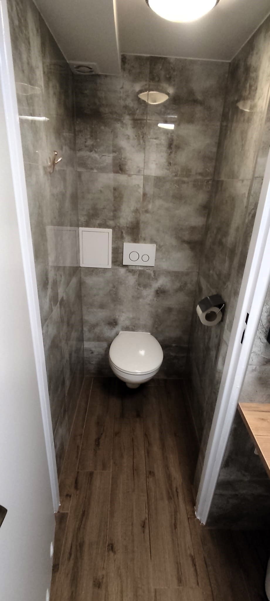 Wąska toaleta wykończona płytkami imitującymi beton na ścianach i drewnopodobną podłogą, z wiszącą muszlą klozetową i minimalistycznym przyciskiem spłukującym.