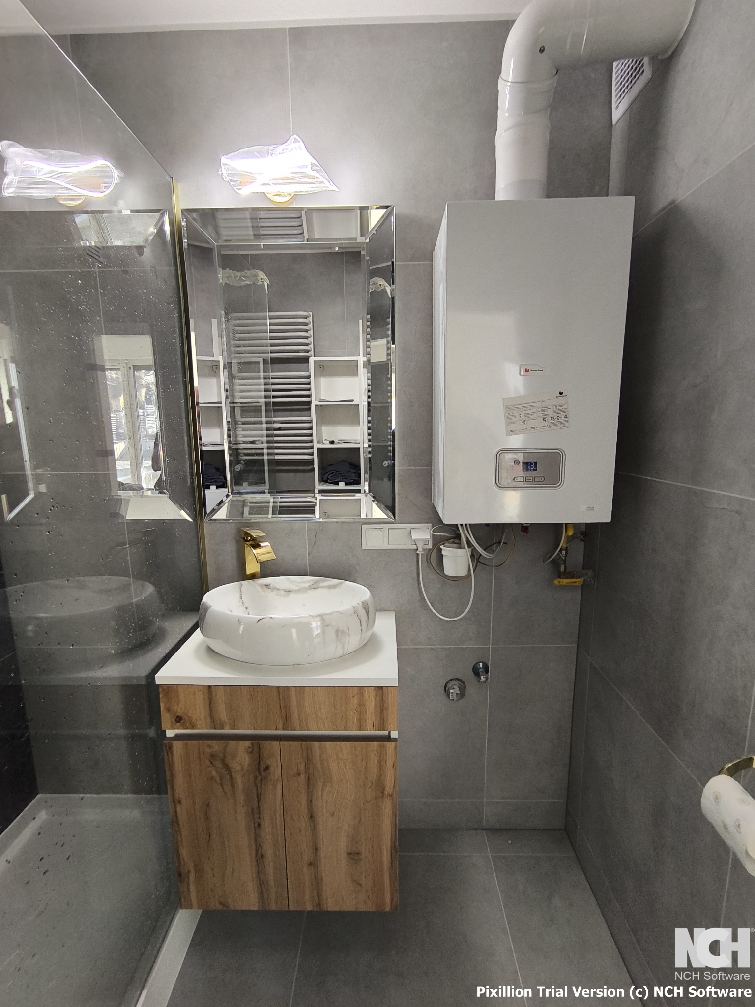 Szara łazienka z marmurową umywalką nablatową, złotą baterią, grzejnikiem drabinkowym odbijającym się w lustrze oraz białym piecem gazowym z widocznymi rurami.