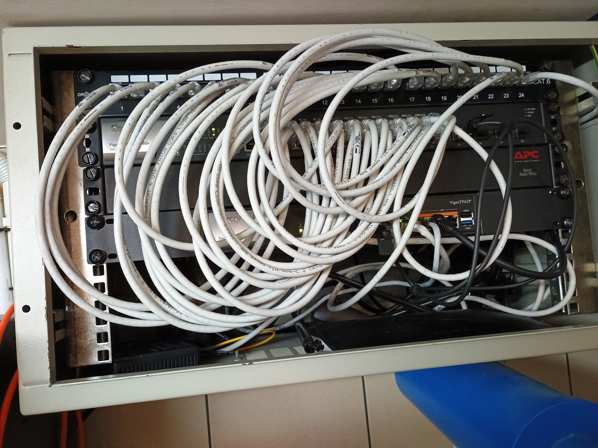 Rozdzielnia sieci LAN w szafie rack z patch panelem, switchem Draytek i listwą zasilającą APC. Widoczne białe kable patchcord CAT6 i przewody zasilające. Niechlujne ułożenie kabli.