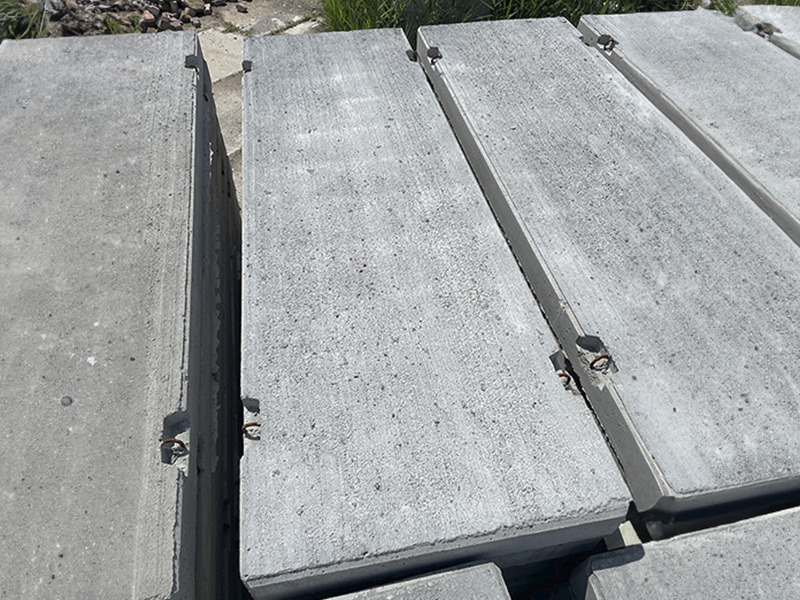 Stos szarych, betonowych płyt z metalowymi uchwytami do podnoszenia, ułożonych jedna na drugiej, widoczne z góry pod lekkim kątem.