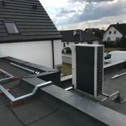 Biała jednostka zewnętrzna pompy ciepła zamontowana na płaskim dachu domu jednorodzinnego, widoczne metalowe wsporniki i elementy instalacji, na tle pochmurne niebo i zabudowa mieszkalna.