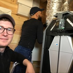 Sanerga - Uśmiechnięty mężczyzna w okularach i czapce pozuje na tle montażu centrali wentylacyjnej z widocznymi filtrami i podłączeniem do kanału wentylacyjnego, drugi mężczyzna w tle pracuje nad instalacją.